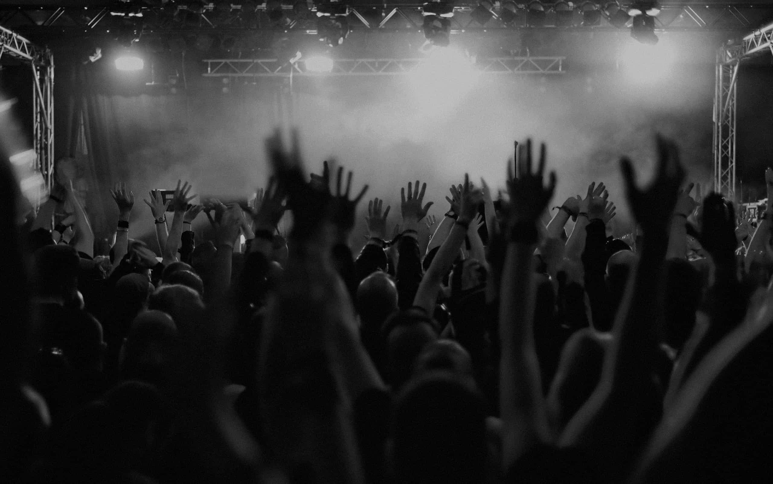 Black-and-white concert crowd raising their hands toward a hazy stage lit by rig lights